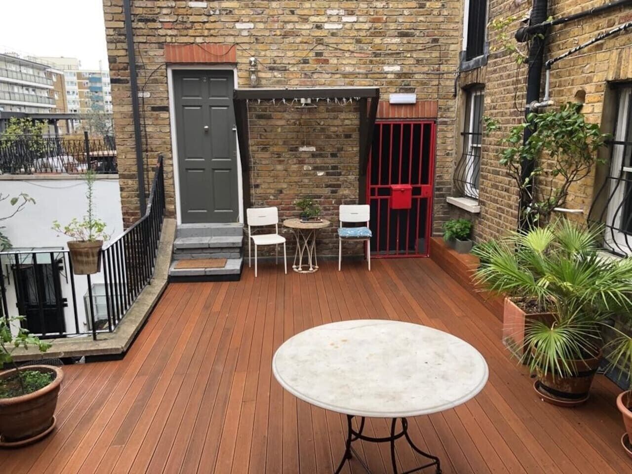 Une terrasse en bois avec une table ronde et deux chaises, un mur en briques, une porte rouge et des plantes en pot.