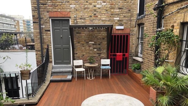 Eine Holzterrasse mit einem runden Tisch und zwei Stühlen, eine Backsteinmauer, ein rotes Tor und Kübelpflanzen.