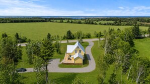 A single-story house with a wrap-around porch, surrounded by a well-maintained lawn and trees, with a road and a body of water in the background. - Cavendish Gables Cottage (Green Gables)