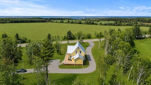 A single-story house with a wrap-around porch, surrounded by a well-maintained lawn and trees, with a road and a body of water in the background. - Cavendish Gables Cottage (Green Gables)
