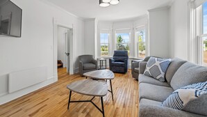 A spacious living room with hardwood flooring, a grey sofa with patterned cushions, a grey armchair, a small wooden coffee table, and a ceiling light fixture. - Cavendish Gables Cottage (Green Gables)