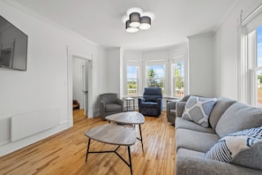 A spacious living room with hardwood flooring, a grey sofa with patterned cushions, a grey armchair, a small wooden coffee table, and a ceiling light fixture. - Cavendish Gables Cottage (Green Gables)