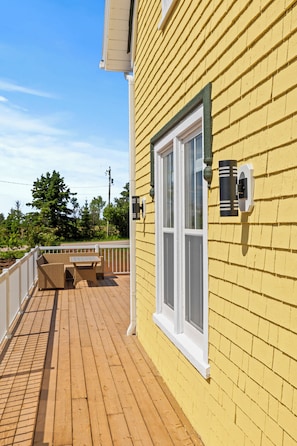 A wooden deck with a white railing, a window with a white frame, and a wall-mounted light fixture. - Cavendish Gables Cottage (Green Gables)