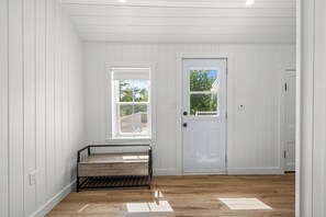 A room with white paneled walls, a wooden floor, a bench, a door, and a window with a view of trees. - Cavendish Gables Cottage (Green Gables)