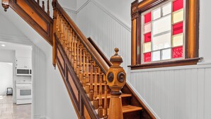 A wooden staircase with a decorative balustrade, a window with red and white panes, and a kitchen area in the background. - Cavendish Gables Cottage (Green Gables)