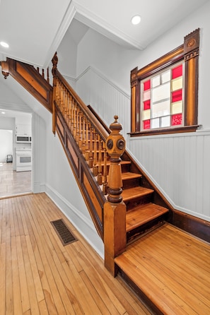 A wooden staircase with a decorative balustrade, a window with red and white panes, and a kitchen area in the background. - Cavendish Gables Cottage (Green Gables)