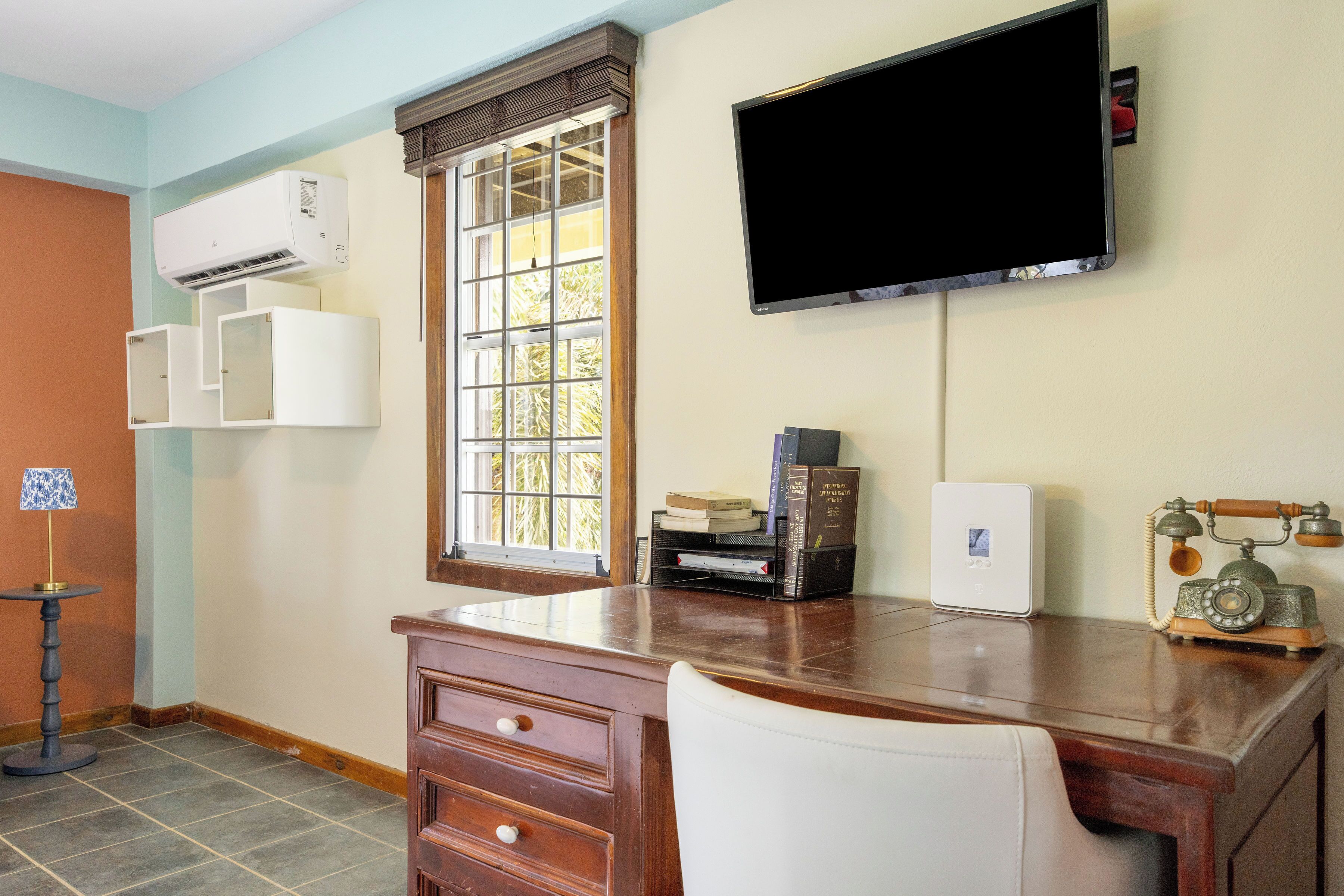 Una habitación con escritorio, una silla, televisor montado en la pared, aire acondicionado y una ventana con persianas.