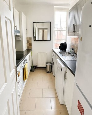 A compact kitchen with white cabinets, a black countertop, a window, and a white refrigerator. - 2-Bedroom Home with FREE Parking near Sydenham Station (Greater London)