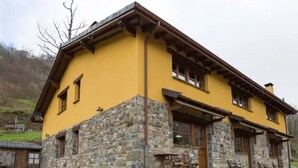 A two-story building with a yellow facade and stone foundation, featuring a porch with outdoor seating. - The LLananzanes Lookout—Asturias Rural—CASA II (Conforcos (Aller))