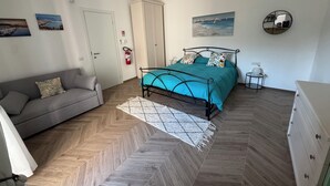 Ein Schlafzimmer mit einem Bett, einem grauen Sofa, einer weißen Kommode und einem Parkettboden mit Zickzackmuster.
