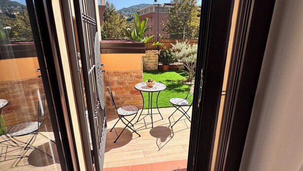 Blick durch eine offene Tür auf eine Terrasse mit Tisch und Stühlen, einen kleinen Garten und ein Gebäude im Hintergrund.