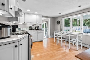 A kitchen with white cabinets, a stove, and a dining area with a table and chairs. - Cozy 2BR Family Home with Fire Pit and Backyard (Barnstable)