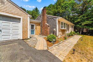 A two-story house with a brick chimney, a garage, and a well-maintained front yard with a pathway. - Cozy 2BR Family Home with Fire Pit and Backyard (Barnstable)
