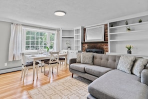A modern living room with a grey sofa, wooden dining table, and a fireplace. - Cozy 2BR Family Home with Fire Pit and Backyard (Barnstable)