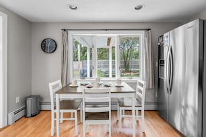 A kitchen with a dining table, chairs, a refrigerator, a clock, and a window with curtains. - Cozy 2BR Family Home with Fire Pit and Backyard (Barnstable)