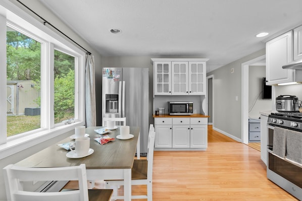 A modern kitchen with white cabinets, a wooden dining table, and a view of the outdoors. - Cozy 2BR Family Home with Fire Pit and Backyard (Barnstable)