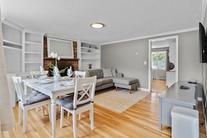 A modern living room with a dining area, a grey sofa, a white table with chairs, and a TV mounted on the wall. - Cozy 2BR Family Home with Fire Pit and Backyard (Barnstable)