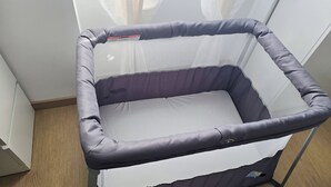 Ein Babyreisebett mit Netzoberteil und gepolsterten Seiten, stehend auf Holzboden, mit weißem Vorhang im Hintergrund.
