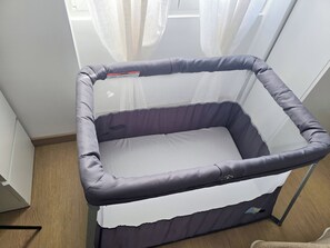 Ein Babyreisebett mit Netzoberteil und gepolsterten Seiten, stehend auf Holzboden, mit weißem Vorhang im Hintergrund.