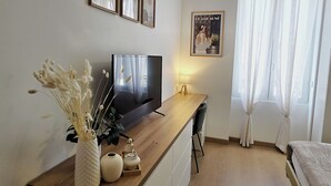 Ein modernes Wohnzimmer mit Flachbildfernseher, Holzschreibtisch, einer Vase mit Trockenblumen und einem gerahmten Poster an der Wand.