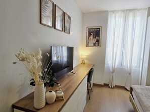 Ein modernes Wohnzimmer mit Flachbildfernseher, Holzschreibtisch, einer Vase mit Trockenblumen und einem gerahmten Poster an der Wand.