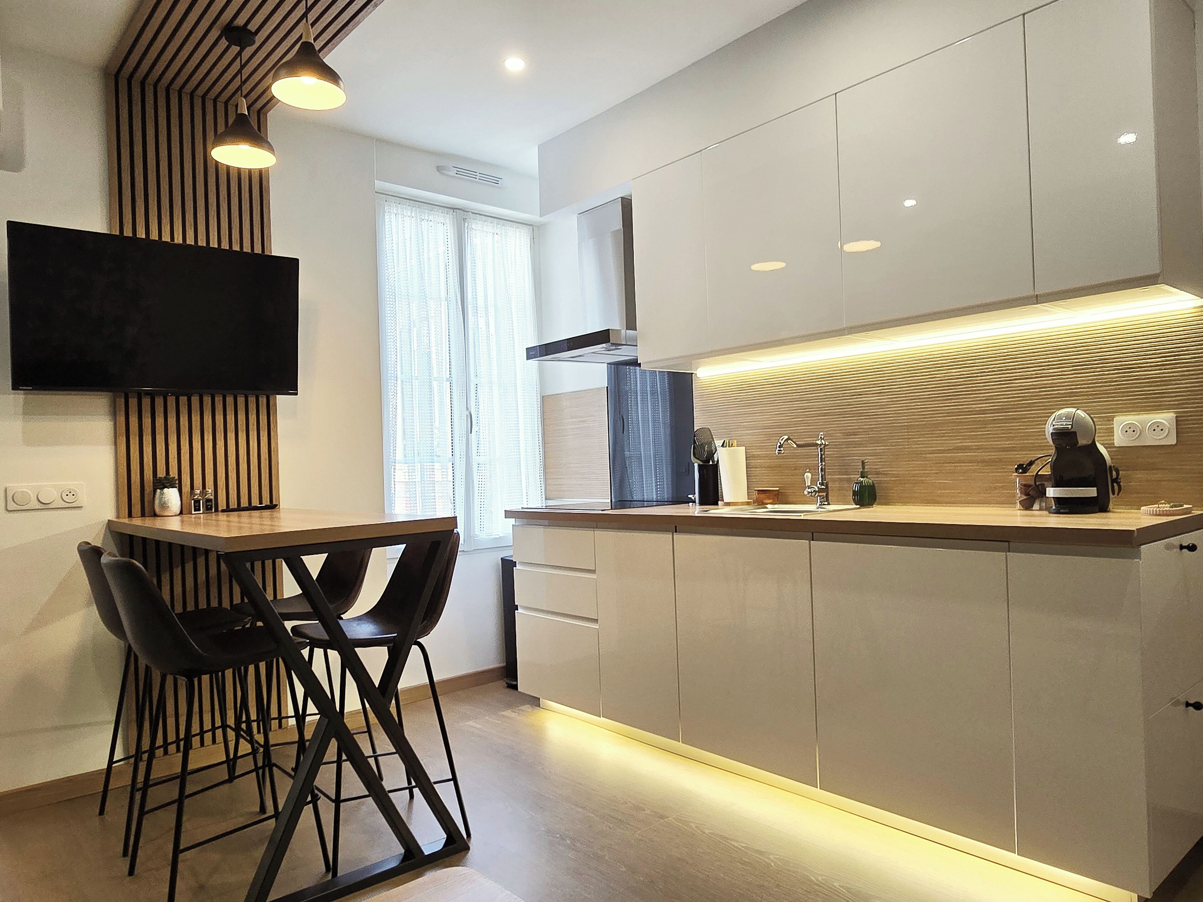 A modern kitchen with a dining area, featuring a flat-screen TV, wooden accents, and a coffee machine.