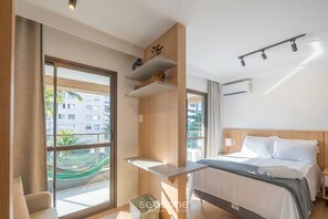 Ein modernes Hotelzimmer mit Bett, einem kleinen Tisch, einem Fenster mit Blick auf Gebäude und einer Hängematte draußen.