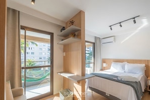 A modern hotel room with a bed, a small table, a window with a view of buildings, and a hammock outside. - Comfort in Condo w/ Pool in Pituba Hpa0202 (Salvador)
