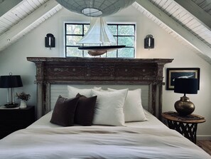 A bedroom with a sloped ceiling, a fireplace, a bed with pillows, a lamp, and a window with a view of trees.