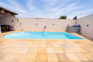A rectangular swimming pool with a tiled deck, a water fountain, and a building with a tiled roof in the background. - Casa com Piscina e Churrasqueira Perto do Centro (Uberlandia)
