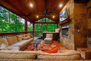 A covered outdoor seating area with a stone fireplace, a television, and a ceiling fan. - Pour Me - Luxury Cabin; Carter Mtn; Sleeps 12 (McCurtain County)