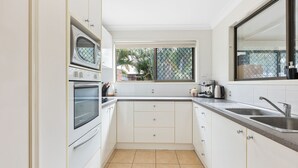 Una cucina compatta con mobili bianchi, forno integrato, macchina da caffè e finestra vista verde.