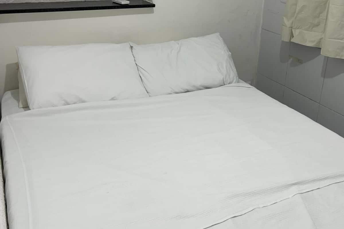 A neatly made bed with white linens in a room with white walls and a towel hanging on the wall.