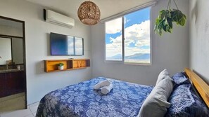 A bedroom with a bed, a TV, a window with a view, and a hanging plant. - Vista Tower, 3-bedroom apartment, panoramic view, rooftop pool and gym (San Salvador)