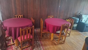 A dining area with two round tables covered with red tablecloths, wooden chairs, and a patterned rug.