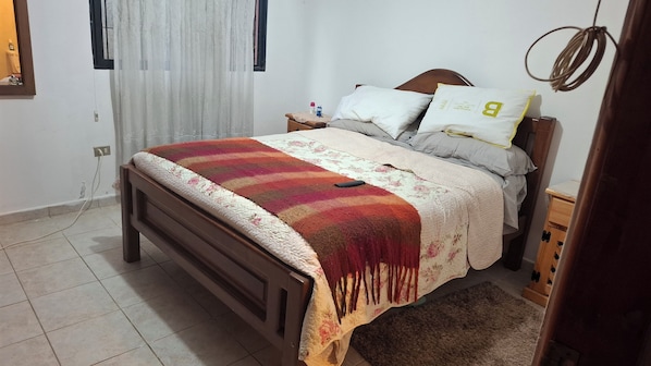 Quarto com cama, mesa de cabeceira, cortinas na janela e tomada na parede.