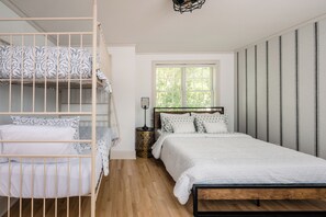 Ein Schlafzimmer mit Etagenbett, einem Einzelbett, einem Nachttisch mit Lampe und einem Fenster mit Blick ins Grüne.
