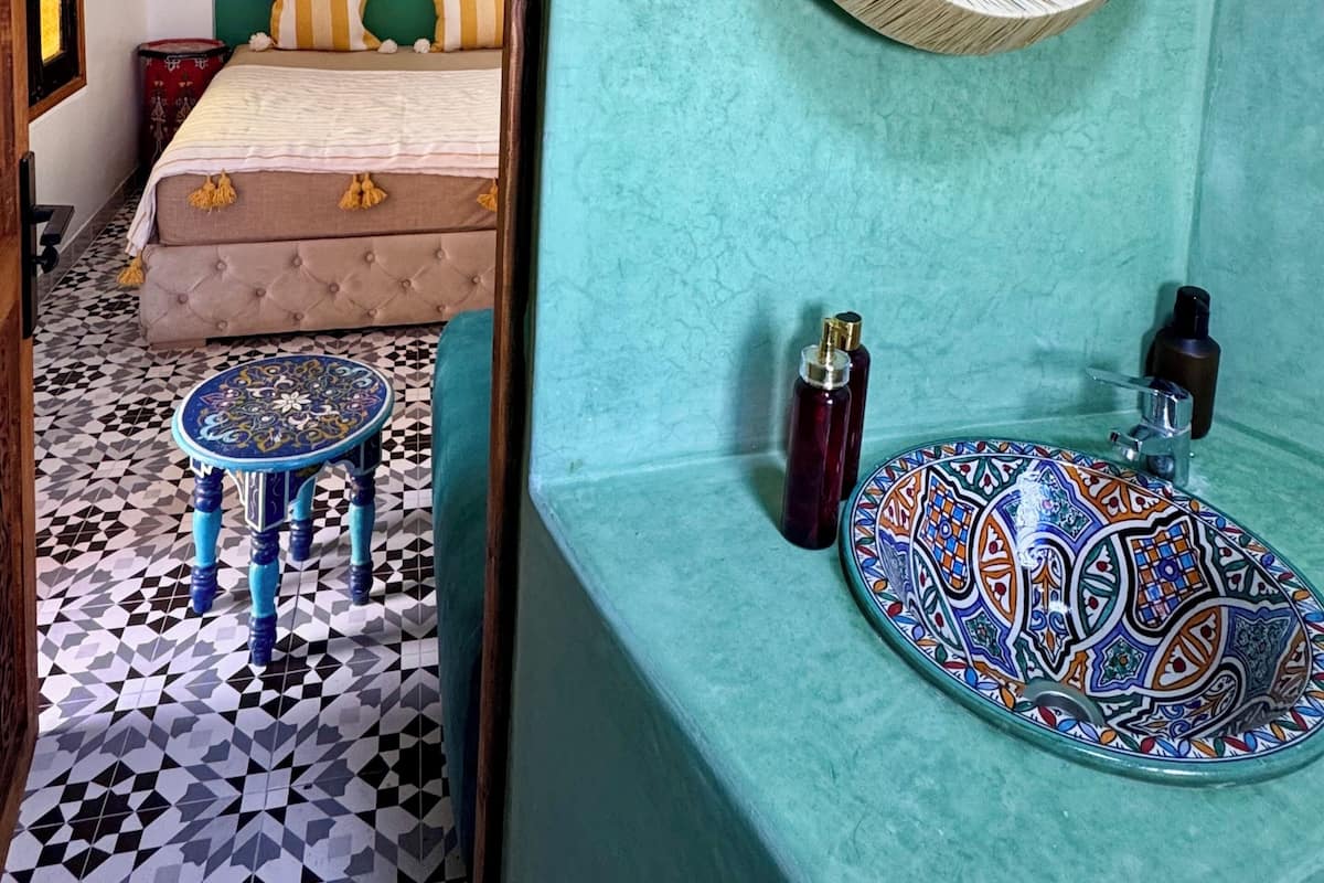 Ein Zimmer mit einem Bett, einem runden Spiegel, einem waschbecken mit Muster und einem kleinen Hocker.