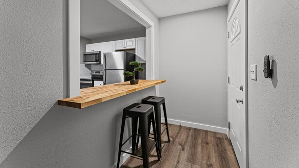A narrow hallway with a kitchen area featuring a wooden bar counter, two black stools, and a potted plant.