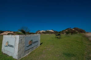 A sign for a 'RESERVA' with a palm tree logo, set against a clear blue sky, with a grassy area and distant hills. - Beach Estates com Piscina Privativa By Carpediem (Aracati)