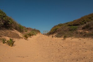 Ein sandiger Pfad führt zu einer Struktur mit blauem Dach, umgeben von spärlicher Vegetation.