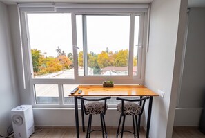 Una habitación con ventana, una mesa de madera, dos sillas, una planta pequeña y un aire acondicionado blanco.