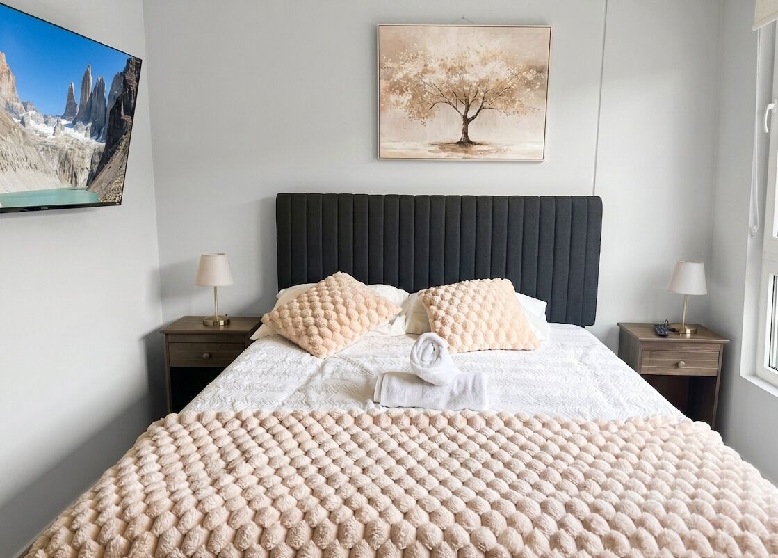 A neatly made bed with a textured blanket, two pillows, a folded towel, and a painting of a tree on the wall.