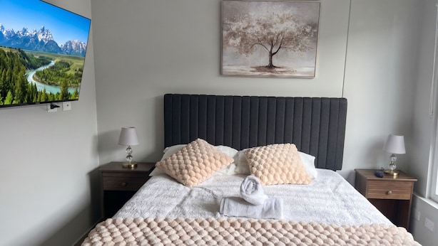 A neatly arranged bedroom with a bed, two bedside tables, a TV mounted on the wall, and a painting above the bed.