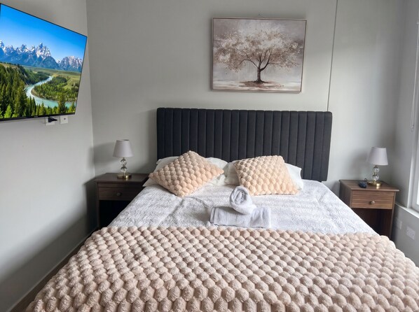 Un dormitorio ordenado con una cama, dos mesitas de noche, un televisor montado en la pared y un cuadro sobre la cama.
