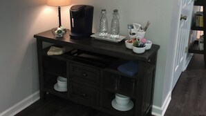 A desk with a lamp, coffee maker, and two glass bottles, a painting on the wall, and a door with a bookshelf.