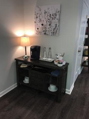 Ein Schreibtisch mit Lampe, Kaffeemaschine und zwei Glasflaschen, ein Bild an der Wand und eine Tür mit Bücherregal.