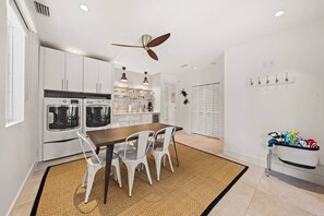 A modern kitchen with white cabinetry, a dining table, chairs, a ceiling fan, and a laundry basket. - Luxurious Retreat At The Whispering Palms (Wabasso Beach)