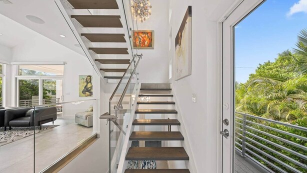 A modern staircase with glass railings, a view of a balcony with greenery, and a living area with a sofa and a painting. - Luxurious Retreat At The Whispering Palms (Wabasso Beach)