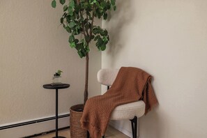 Ein Zimmer mit einem weißen Stuhl, einer braunen Tagesdecke, einem kleinen Tisch mit einer Pflanze und einem Blumentopf.
